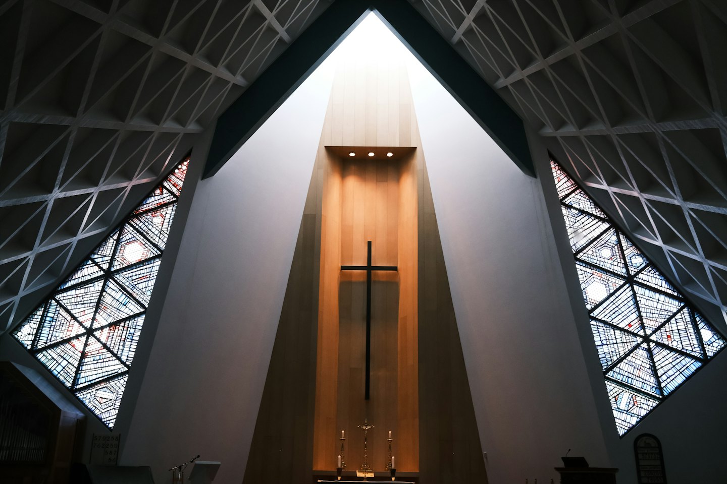 A tall dark cross stands against a beige wooden wall in a modern chapel.